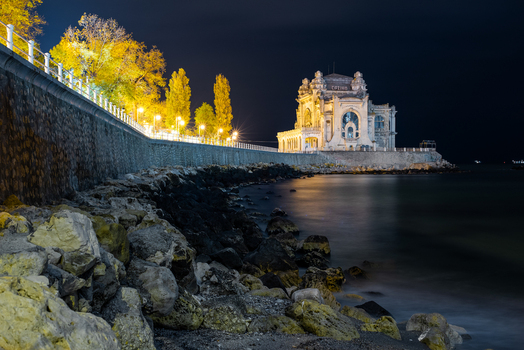 View of the illuminated Casino of Constanta at night featuring a rocky shoreline coastal promenade with trees and calm sea in Constanta Judetul Constanta Romania. View of the illuminated Casino of Constanta at night featuring a rocky shoreline coastal promenade with trees and calm sea in Constanta Judetul Constanta Romania.
