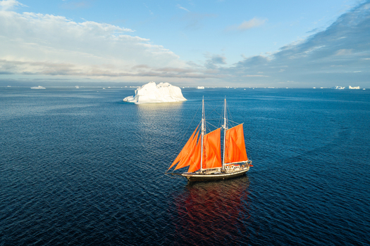 Aerial view of a sailboat with vibrant orange sails navigating the deep blue waters near a large white iceberg in Greenland. Aerial view of a sailboat with vibrant orange sails navigating the deep blue waters near a large white iceberg in Greenland.