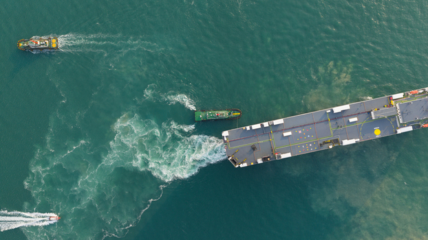 Aerial view of a large cargo ship being assisted by tugboats in the turquoise waters of the port in Laem Chabang Chon Buri Thailand.
