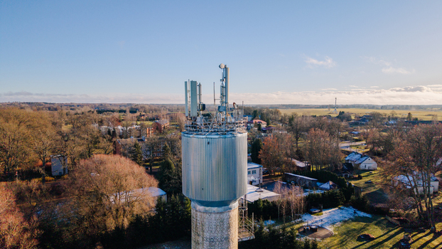 Valmiera Latvia 11 November 2025 Aerial view of a telecommunications tower with antennas and satellite dishes rising above a rural landscape with trees and buildings under a clear sky.
