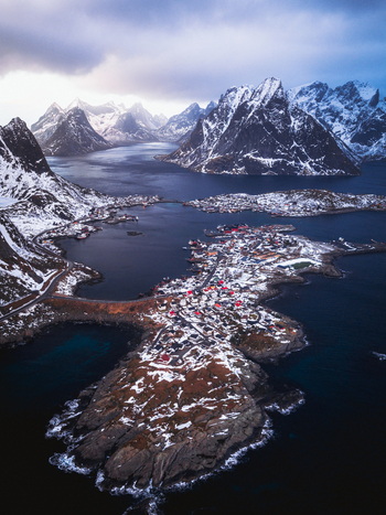 Aerial view of snow dusted rooftops nestled amid the jagged snow capped peaks rising majestically from the icy waters Reinebringen Nordland Norway.