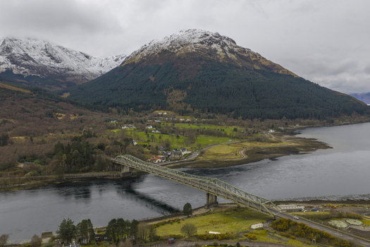 Aerial view of Ballachulish Bridge spanning Loch Leven with snow capped mountains and lush green hills under an overcast sky in Ballachulish Scotland United Kingdom.