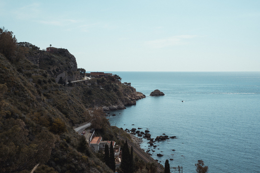 View of rugged cliffs meet the tranquil sea with winding roads hugging the coastline under a clear sky creating a serene coastal scene Taormina Sicily Italy. View of rugged cliffs meet the tranquil sea with winding roads hugging the coastline under a clear sky creating a serene coastal scene Taormina Sicily Italy.