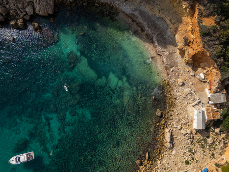 Aerial view of Cala Llebeig with its turquoise waters rocky coastline small white stone buildings a boat and a person on a paddleboard in El Poble Nou de Benitatxell Valencian Community Spain.