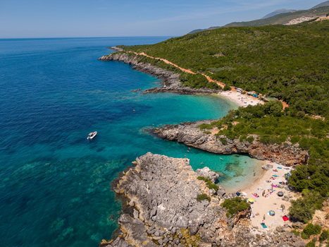 Aerial view of Aquarium Beach with turquoise water rocky cliffs and people on the sandy shore surrounded by lush green hills in Jala Vlore County Albania.