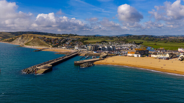 Aerial view of West Bay featuring the harbour entrance sandy beach and coastal cliffs under a blue sky with clouds in West Bay England United Kingdom. Aerial view of West Bay featuring the harbour entrance sandy beach and coastal cliffs under a blue sky with clouds in West Bay England United Kingdom.