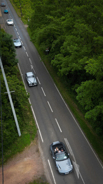 Aerial view of luxury and classic cars with people in a convertible driving along a paved road surrounded by trees in La Chapelle de Guinchay Bourgogne Franche Comte France.