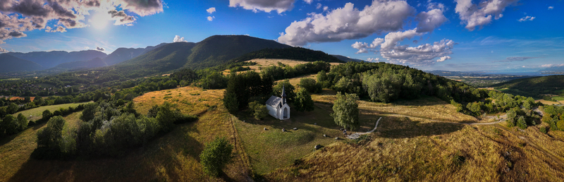 Aerial view of Chapelle Notre Dame de Riantmont a small white chapel on a grassy hill surrounded by lush green trees and rolling mountains under a blue sky in Vesancy Auvergne Rhone Alpes France.