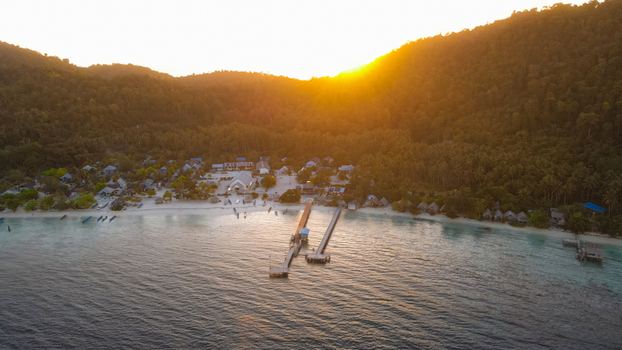 Aerial view of a coastal village with wooden piers extending into the sea lush green hills and golden hour sunlight in Raja Ampat Southwest Papua Indonesia.