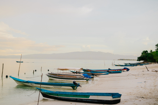 View of traditional wooden outrigger boats moored on a serene white sand beach with calm turquoise waters and distant mountains under a soft sky in Raja Ampat Southwest Papua Indonesia.
