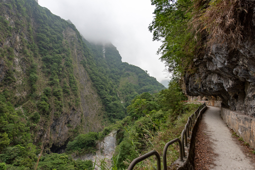 View of the narrow hiking trail carved into the steep rocky cliffs of Taroko National Park with lush green mountains and a metal railing in Xiulin Township Hualien County Taiwan.