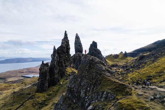 Aerial view of The Storr with its iconic rock formations and a person in a red jacket standing on the peak surrounded by grassy slopes and the sea in Portree Scotland United Kingdom. Aerial view of The Storr with its iconic rock formations and a person in a red jacket standing on the peak surrounded by grassy slopes and the sea in Portree Scotland United Kingdom.