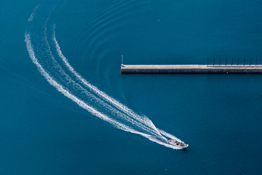 Aerial view of a small motorboat leaving a curved white wake on the deep blue sea surface near a concrete pier at the Itoshima Peninsula Itoshima Fukuoka Japan. Aerial view of a small motorboat leaving a curved white wake on the deep blue sea surface near a concrete pier at the Itoshima Peninsula Itoshima Fukuoka Japan.