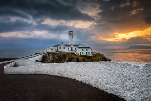 View of Fanad Head Lighthouse on a rocky cliff under a dramatic sunset sky with a white stone wall in the foreground Baile Lair County Donegal Ireland.