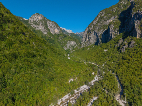 Aerial view of the lush green valley of Theth surrounded by rocky mountains with a winding riverbed and dense forest under a clear blue sky in Theth Shkoder County Albania. Aerial view of the lush green valley of Theth surrounded by rocky mountains with a winding riverbed and dense forest under a clear blue sky in Theth Shkoder County Albania.
