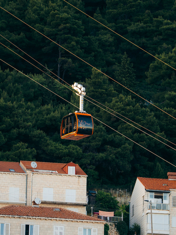 View of orange Dubrovnik Cable Car cabin ascending over traditional stone houses with red tiled roofs against a dense green forest in Dubrovnik Dubrovnik Neretva County Croatia. View of orange Dubrovnik Cable Car cabin ascending over traditional stone houses with red tiled roofs against a dense green forest in Dubrovnik Dubrovnik Neretva County Croatia.