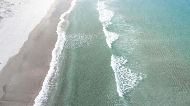 Aerial view of the pristine white sand beach and clear turquoise water with gentle waves rolling onto the shore in Omaha Auckland New Zealand.