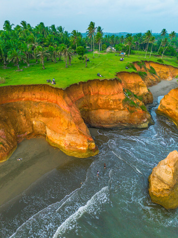 Aerial view of dramatic orange cliffs and lush green palm trees overlooking the sandy beach and ocean waves in Bali Indonesia. Aerial view of dramatic orange cliffs and lush green palm trees overlooking the sandy beach and ocean waves in Bali Indonesia.