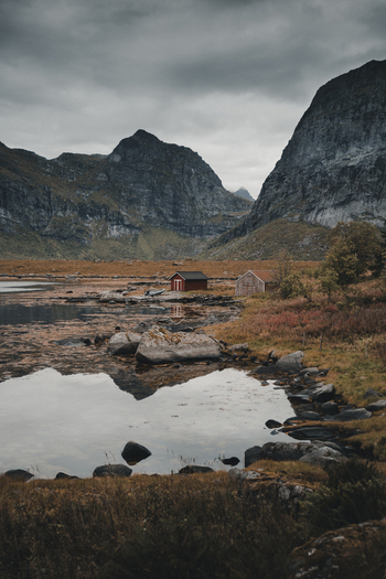Nordland Norway 09 September 2022 View of red wooden cabins nestled by a calm lake surrounded by dramatic steep rocky mountains in the Lofoten islands under a cloudy sky. Nordland Norway 09 September 2022 View of red wooden cabins nestled by a calm lake surrounded by dramatic steep rocky mountains in the Lofoten islands under a cloudy sky.