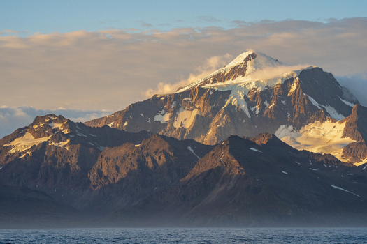 View of snow capped mountain peaks and glaciers illuminated by golden hour light rising above the sea in South Georgia and the South Sandwich Islands.