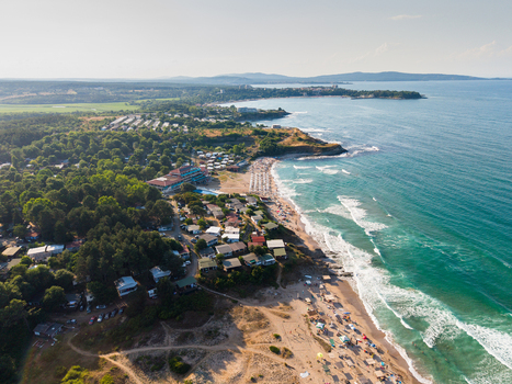 Aerial view of Camping Yug situated along the sandy coastline with small cabins lush green trees and turquoise sea waves in Kiten Burgas Bulgaria.