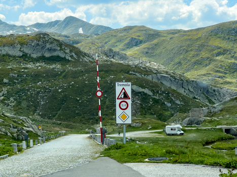 View of the historic Tremola road at Sankt Gotthard Pass with a road sign and barrier against a backdrop of rugged mountains and green slopes Airolo Ticino Switzerland.