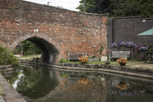 View of a historic red brick arch bridge over a calm canal with wooden benches and colorful potted flowers along the towpath in Coventry England United Kingdom.