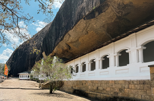 View of Dambulla Cave Temple with its white colonnade built into the massive rock face under a clear blue sky in Sri Lanka.
