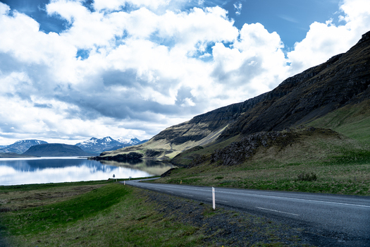 View of the winding coastal road along Hvalfjordur fjord with steep mountains and calm water under a cloudy sky in Iceland. View of the winding coastal road along Hvalfjordur fjord with steep mountains and calm water under a cloudy sky in Iceland.