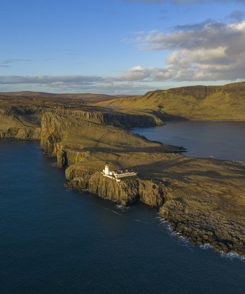 Aerial view of Neist Point Lighthouse on the edge of dramatic rocky cliffs overlooking the blue sea under a soft evening sky Isle of Skye Scotland United Kingdom. Aerial view of Neist Point Lighthouse on the edge of dramatic rocky cliffs overlooking the blue sea under a soft evening sky Isle of Skye Scotland United Kingdom.