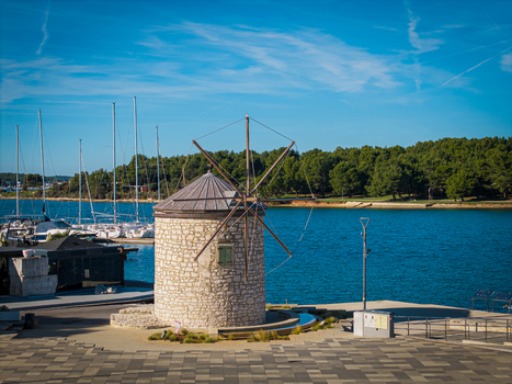 Aerial view of the stone windmill standing proudly by the azure sea with masts of sailboats piercing the horizon and lush trees lining the coast Medulin Istria County Croatia.