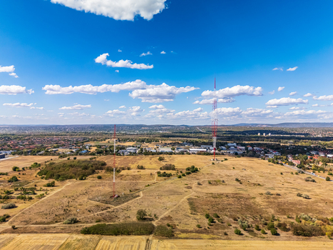 Aerial view of two tall radio transmission towers standing in a dry grassy field with distant town buildings under a clear blue sky in Szigetszentmiklos Pest County Hungary. Aerial view of two tall radio transmission towers standing in a dry grassy field with distant town buildings under a clear blue sky in Szigetszentmiklos Pest County Hungary.