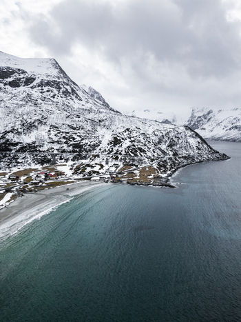 Aerial view of the snowy mountains and turquoise waters of Grotfjord with a sandy beach and coastal village in Tromvik Troms Norway. Aerial view of the snowy mountains and turquoise waters of Grotfjord with a sandy beach and coastal village in Tromvik Troms Norway.