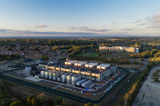Aerial view of a modern data center facility featuring rooftop cooling units and a secure fenced perimeter situated amidst a suburban landscape during the golden hour United Kingdom. Aerial view of a modern data center facility featuring rooftop cooling units and a secure fenced perimeter situated amidst a suburban landscape during the golden hour United Kingdom.