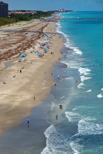 Aerial view of the sandy shoreline with people sunbathing under umbrellas turquoise ocean waves and the Juno Beach Pier in Juno Beach Florida United States. Aerial view of the sandy shoreline with people sunbathing under umbrellas turquoise ocean waves and the Juno Beach Pier in Juno Beach Florida United States.