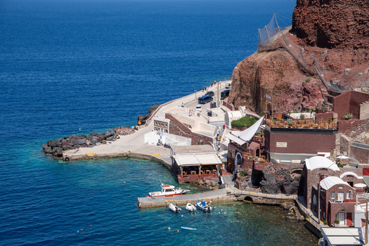 View of the Old Port of Fira with small boats moored at the pier beneath steep red volcanic cliffs and the turquoise Aegean Sea in Fira Greece. View of the Old Port of Fira with small boats moored at the pier beneath steep red volcanic cliffs and the turquoise Aegean Sea in Fira Greece.