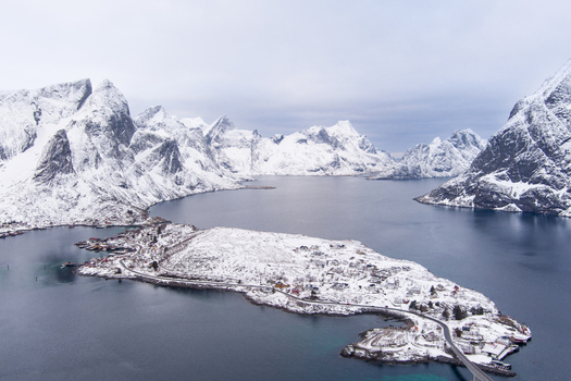 Aerial view of the snow covered village of Reine in the Lofoten islands featuring traditional houses winding roads and dramatic mountain peaks rising from the dark sea in Nordland Norway.
