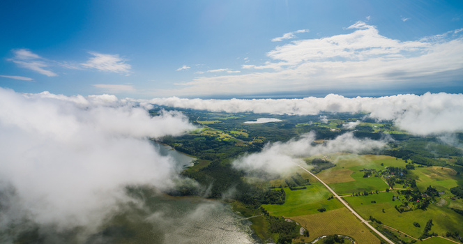 Aerial view of the lush green countryside featuring rolling fields a winding road and a lake partially obscured by low hanging white clouds Latvia. Aerial view of the lush green countryside featuring rolling fields a winding road and a lake partially obscured by low hanging white clouds Latvia.