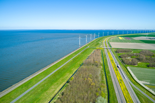 Aerial view of wind turbines along the coastline and a highway running through green fields in the Netherlands.