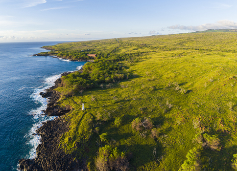 Aerial view of Mahukona Beach Park featuring the rugged volcanic coastline a small white lighthouse and lush green hills under a clear sky in Waimea Hawaii United States.
