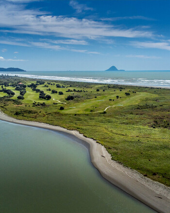 Aerial view of a coastal golf course nestled between sandy beach and green dunes with the Pacific Ocean and a distant island under a blue sky in Ohope Bay of Plenty Region New Zealand.