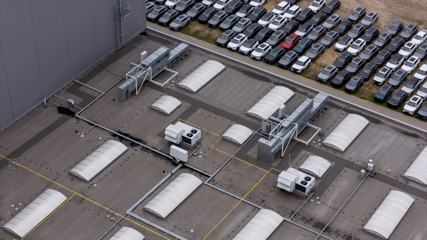 Aerial view of a large industrial factory roof featuring multiple skylights and HVAC units with a parking lot filled with numerous new cars parked in rows in Gdansk Poland. Aerial view of a large industrial factory roof featuring multiple skylights and HVAC units with a parking lot filled with numerous new cars parked in rows in Gdansk Poland.
