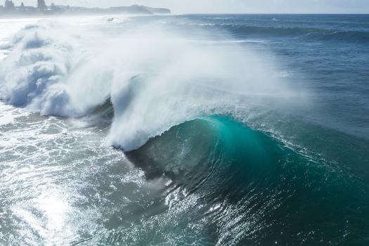 Aerial view of a powerful turquoise ocean wave breaking with white sea spray and foam near the coastline in Narrabeen New South Wales Australia.