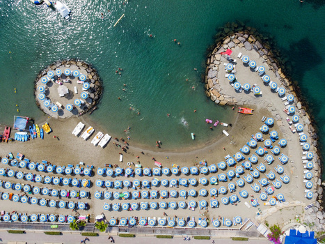 Aerial view of rows of blue and white umbrellas on a sandy beach with people swimming and relaxing near stone breakwaters San Lorenzo Liguria Italy.
