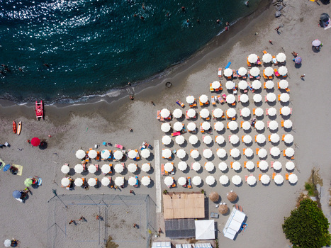 Aerial view of organized rows of white beach umbrellas and sunbeds on the sandy shore with people swimming in the clear blue sea San Lorenzo Liguria Italy.