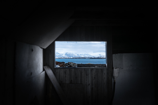 View of abandoned farmhouse interior looking out towards snowy mountains and the sea through a wooden window frame Hafnarfjordur Hafnarfjardarkaupstad Iceland. View of abandoned farmhouse interior looking out towards snowy mountains and the sea through a wooden window frame Hafnarfjordur Hafnarfjardarkaupstad Iceland.