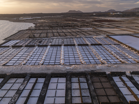 Aerial view of Salinas de Janubio featuring geometric salt pans volcanic soil borders and reflective water pools under a warm sunset sky in Las Palmas Canarias Spain. Aerial view of Salinas de Janubio featuring geometric salt pans volcanic soil borders and reflective water pools under a warm sunset sky in Las Palmas Canarias Spain.