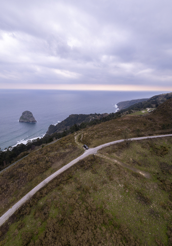 Aerial view of the rocky islet of San Juan de Gaztelugatxe with a camper van parked on a winding coastal road overlooking the sea in Gaztelugatxeko Doniene Euskadi Spain. Aerial view of the rocky islet of San Juan de Gaztelugatxe with a camper van parked on a winding coastal road overlooking the sea in Gaztelugatxeko Doniene Euskadi Spain.