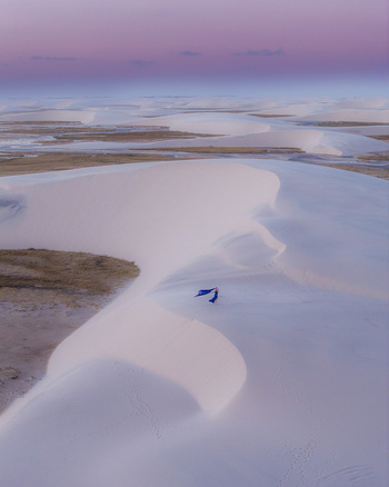 Aerial view of Parque Nacional dos Lencois Maranhenses with a woman in a blue dress standing on white sand dunes under a purple sunset sky in Lencois Maranhenses MA Brazil. Aerial view of Parque Nacional dos Lencois Maranhenses with a woman in a blue dress standing on white sand dunes under a purple sunset sky in Lencois Maranhenses MA Brazil.
