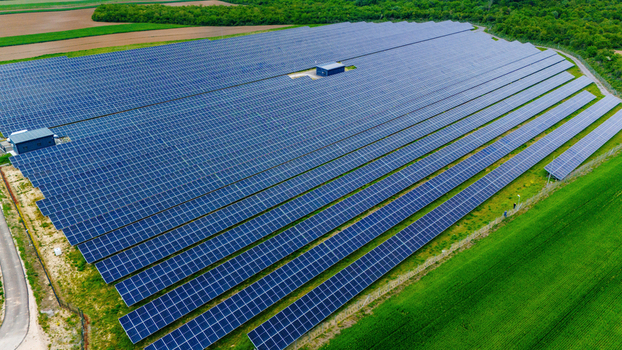 Aerial view of a large solar farm with rows of blue photovoltaic panels surrounded by lush green fields and dense forest in Sabac Vojvodina Serbia.